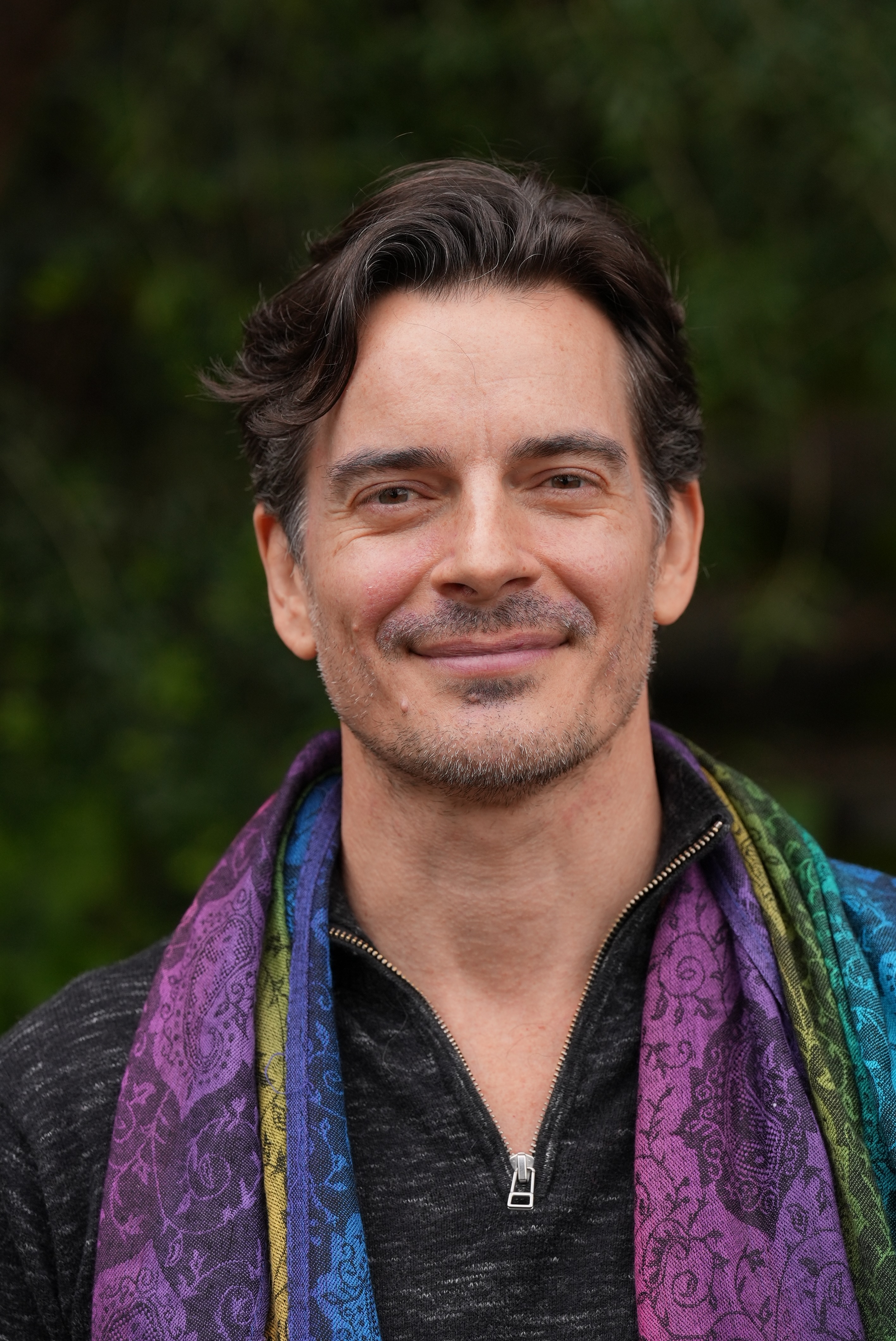 Portrait of Romie Kind: head and shoulders, outdoors against green foliage, dark-grey quarter-zip with a paisley scarf in purple, teal, and chartreuse, warm direct expression.