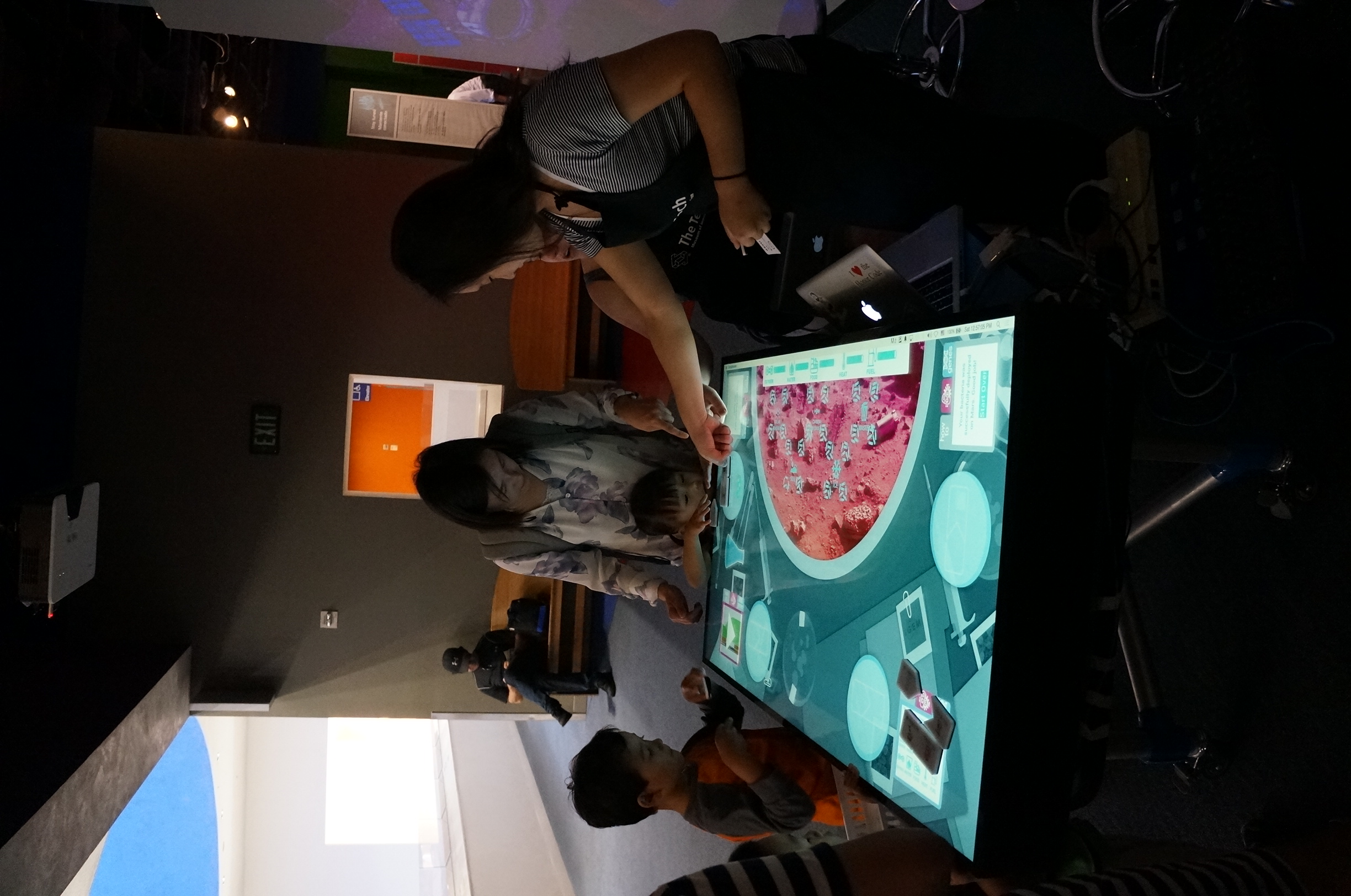 Romie holding an early BacPack prototype, a wooden tabletop unit marked with orange BIOHACK tape, gesturing toward the camera. A monitor in the background shows a CAD wireframe of the same enclosure.