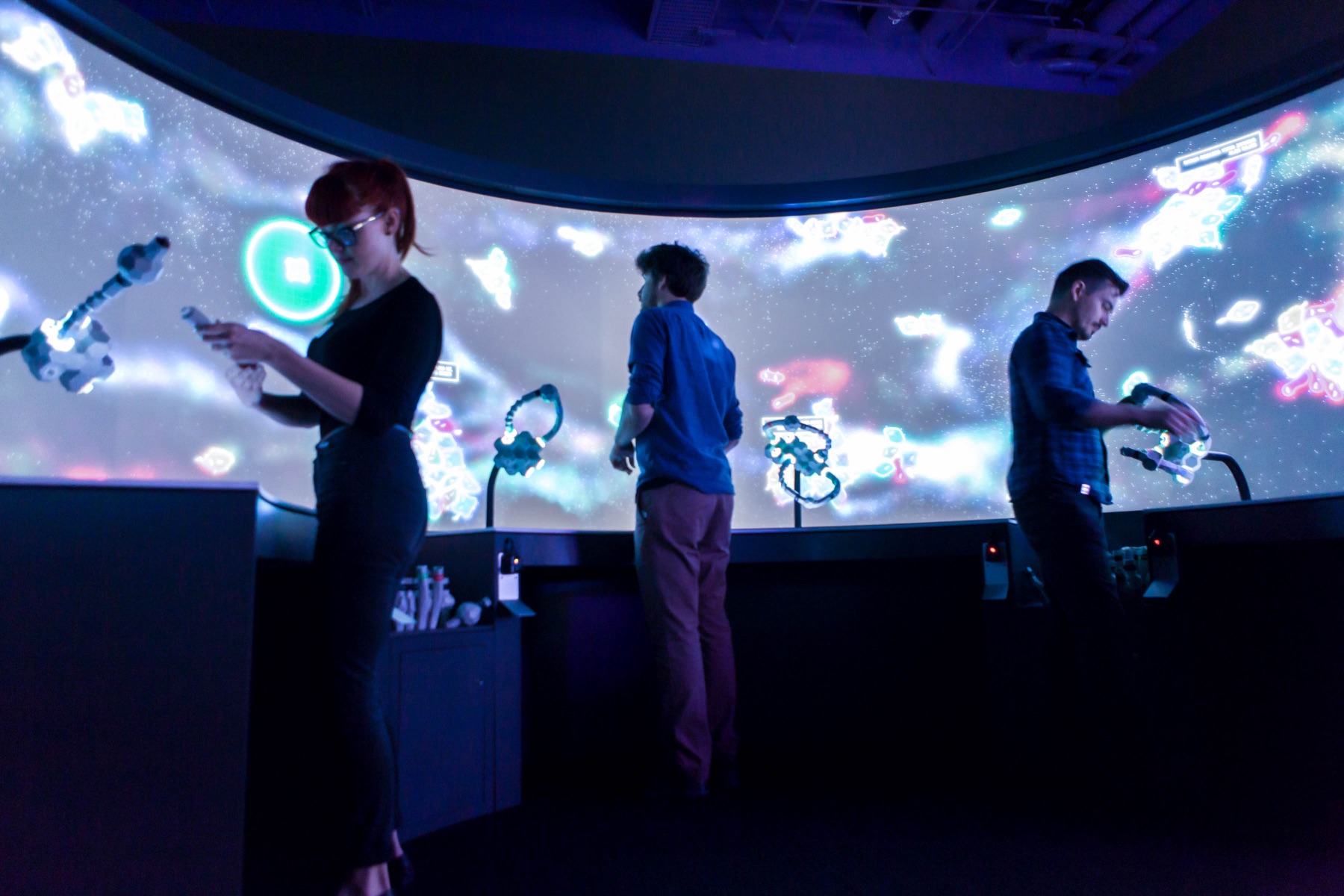 Three visitors stand at the curved BioPool projection wall, each holding a snake-arm tangible controller. Glowing synthetic organisms drift across the wide screen in front of them.