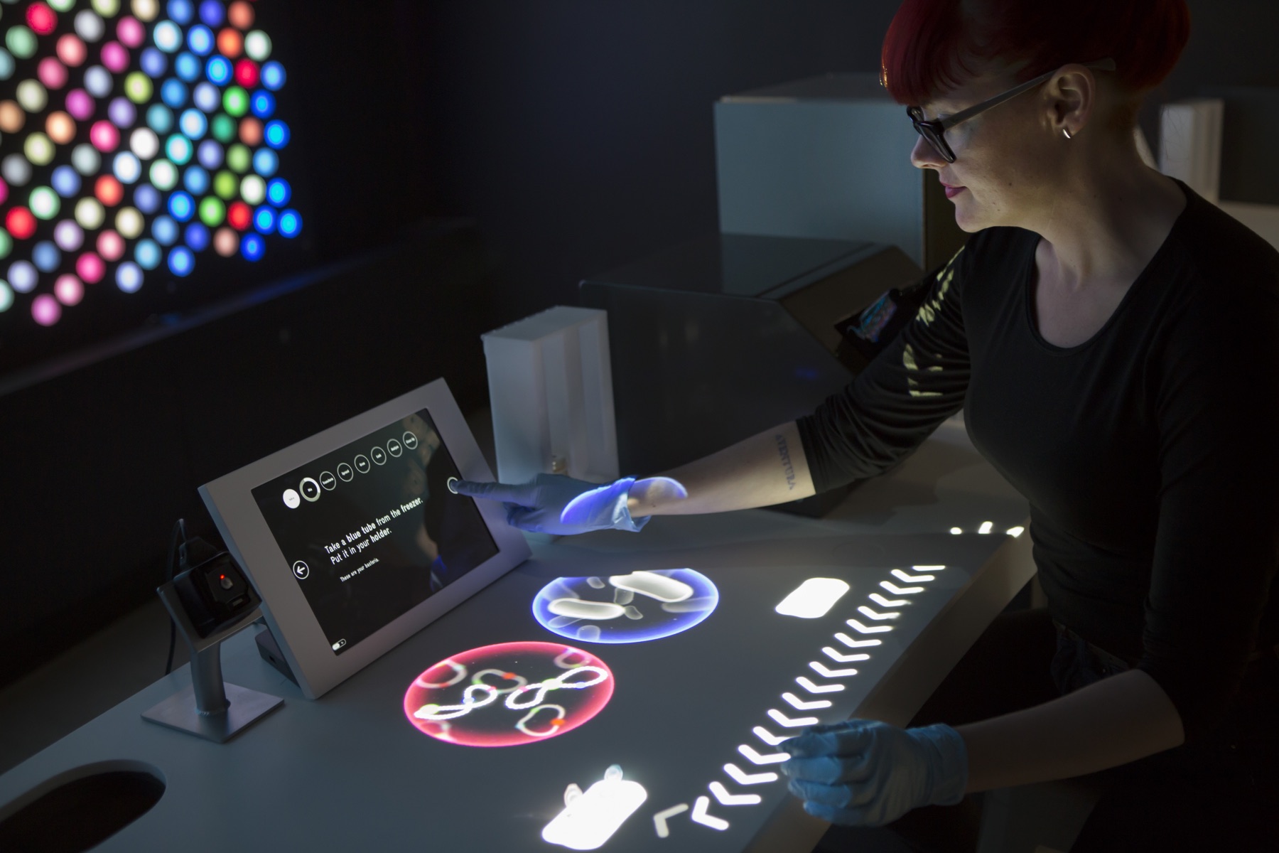 A researcher in a black top and blue nitrile gloves works at the Living Colors Lab tangible table in BioDesign Studio, moving labware tokens. A wall of small multicolored lights glows in the dark background.