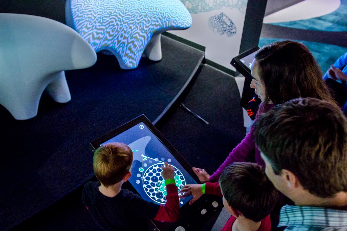 Three young visitors and an adult lean over an interactive tabletop in BioDesign Studio at The Tech Interactive. The screen shows a circular cell-design interface; in the background, an organic-form sculpture printed with a generative pattern is partly visible.