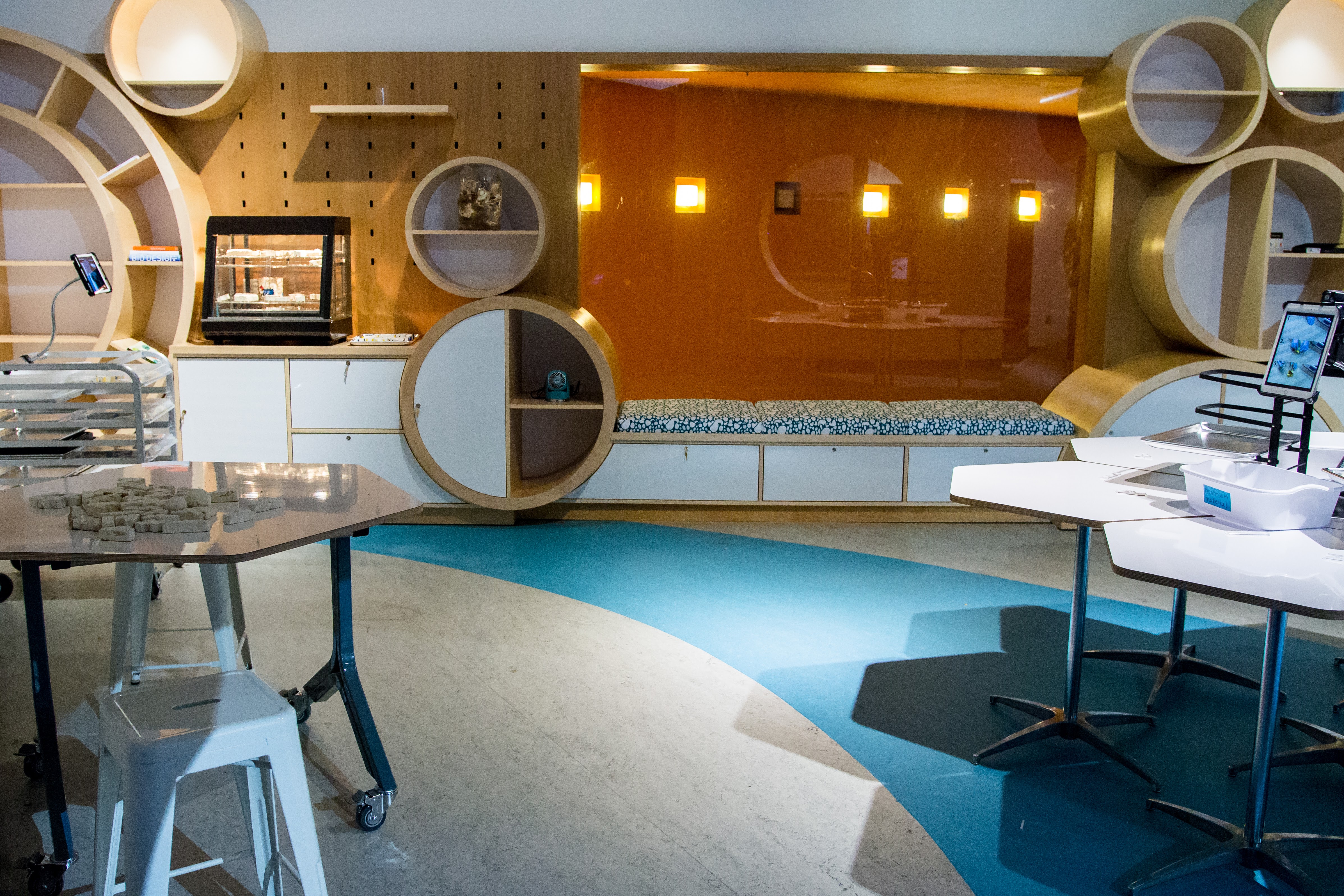An empty BioTinkering Lab interior at The Tech Interactive: warm wood-toned cabinetry with circular display cutouts, a backlit yellow-orange feature panel, hexagonal-base workbenches, and stools arranged for visitor activities. A teal-and-cream patterned floor sweeps through the space.