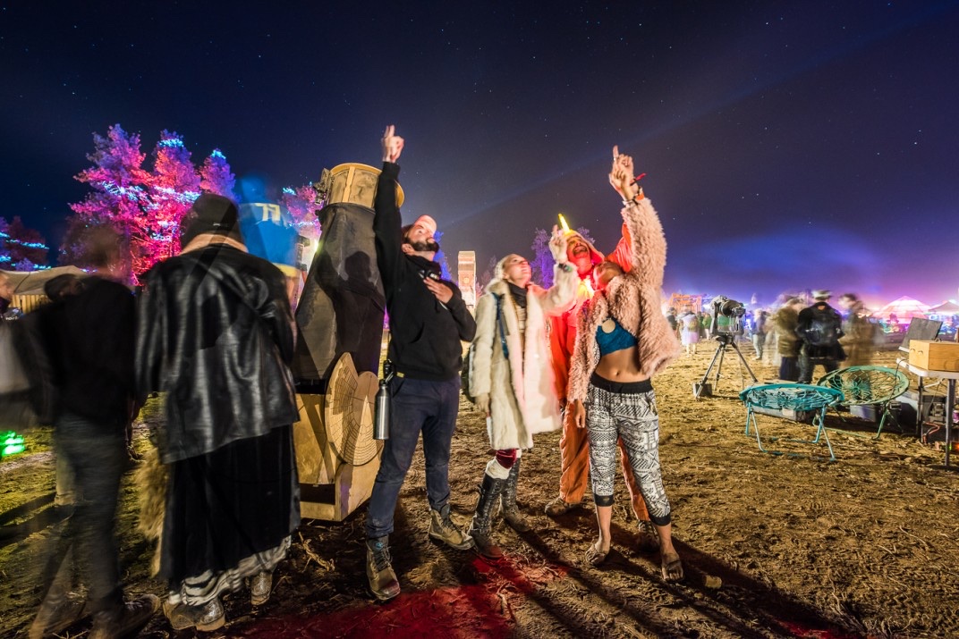 Nighttime at the 2017 Oregon Eclipse Festival. People gather around a tall carved totem under a star-filled sky, with colored lights glowing on trees in the background.