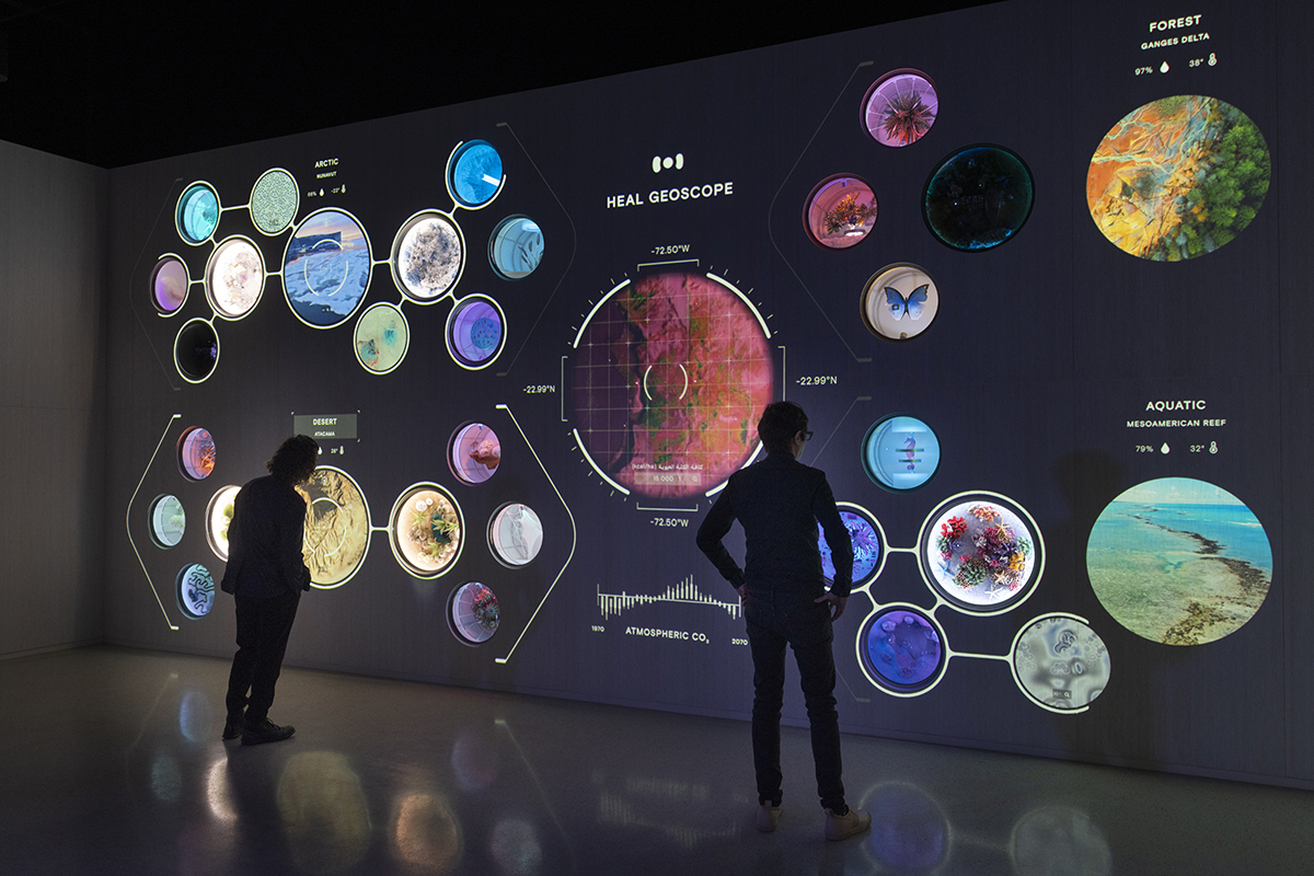 Two figures silhouetted against the HEAL Geoscope wall at the HEAL Institute pavilion. The wall shows a hexagonal grid of circular ecology displays for Arctic, Forest (Ganges Delta), and Aquatic (Mesoamerican Reef) zones, with a central radial visualization of atmospheric CO₂.