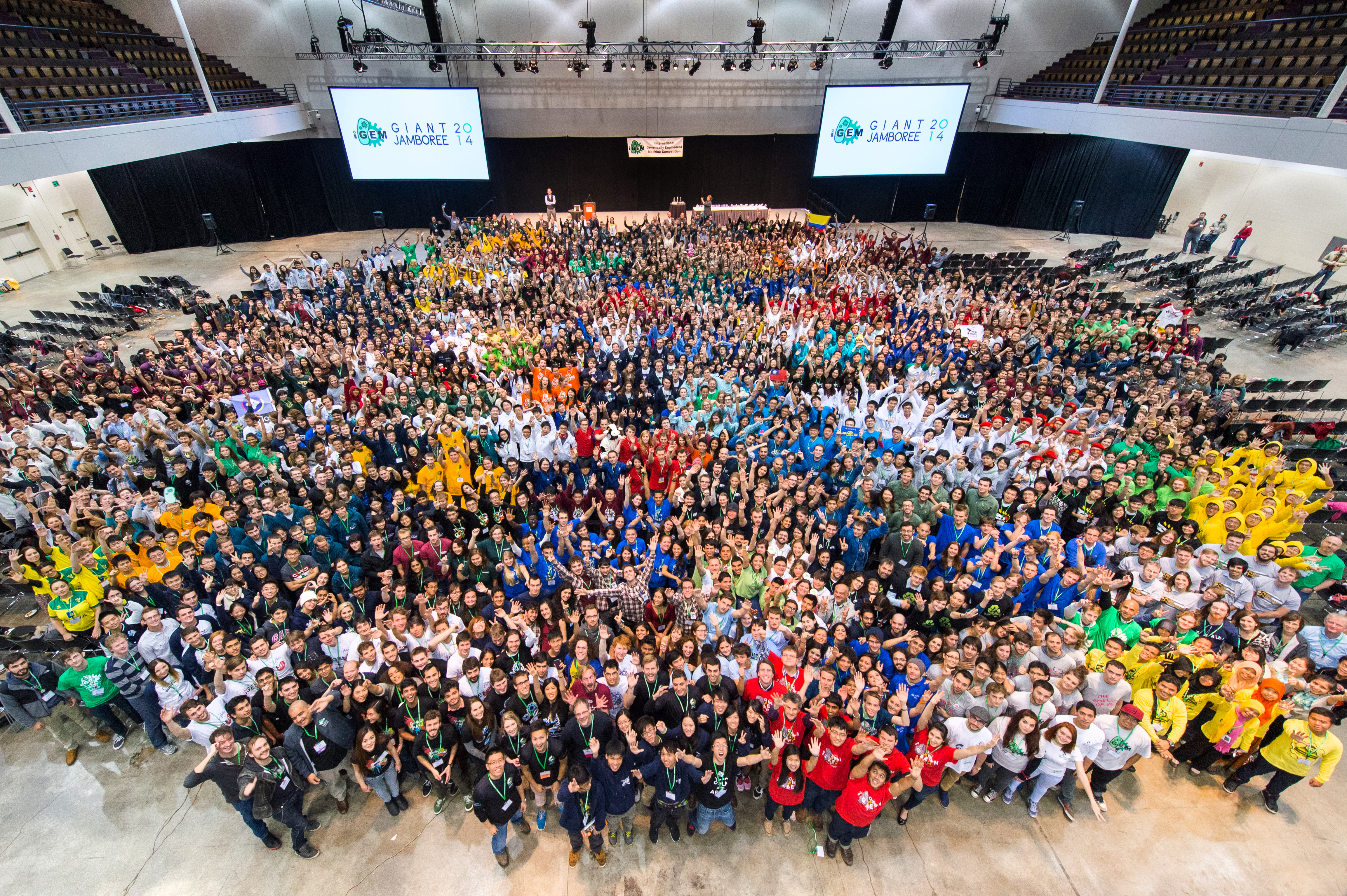 Aerial view of the iGEM Giant Jamboree floor, packed with hundreds of student teams in coordinated team-color clothing arranged in a vast curved formation under stage screens reading GIANT JAMBOREE.