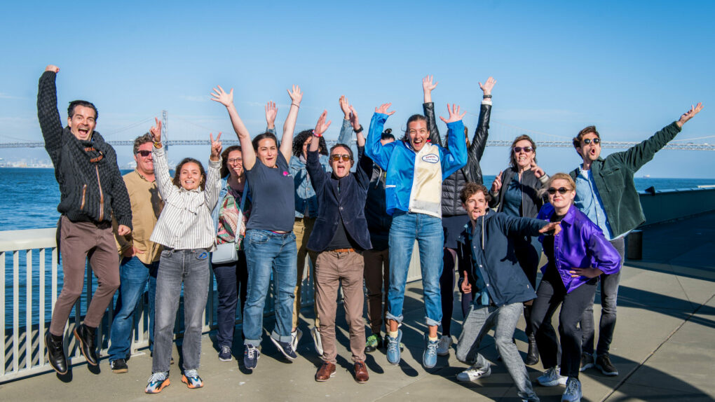 About a dozen bootcamp participants jump with arms raised on a waterfront terrace, the San Francisco Bay Bridge spanning the water behind them under a clear sky.
